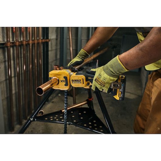 A person wearing green work gloves using a yellow DEWALT cordless power tool to cut a copper pipe, with the tool fixed on a stand in a workshop.