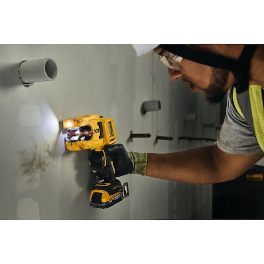 A person wearing safety gear uses a yellow DEWALT cordless tool to cut into a wall with visible pipes.