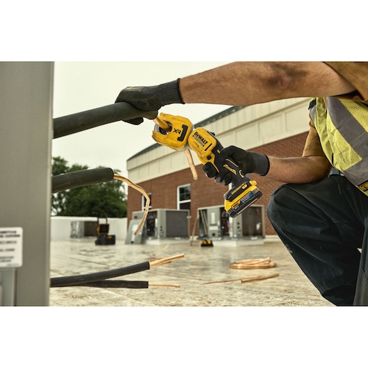 A worker using a DEWALT cordless power tool to cut thick copper cables outdoors at a construction or industrial site. The worker is wearing gloves and safety gear.