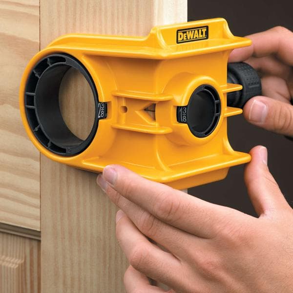 Uma pessoa está usando um kit amarelo para instalação de fechadura de porta da DEWALT para alinhar e marcar um furo em uma moldura de porta de madeira.