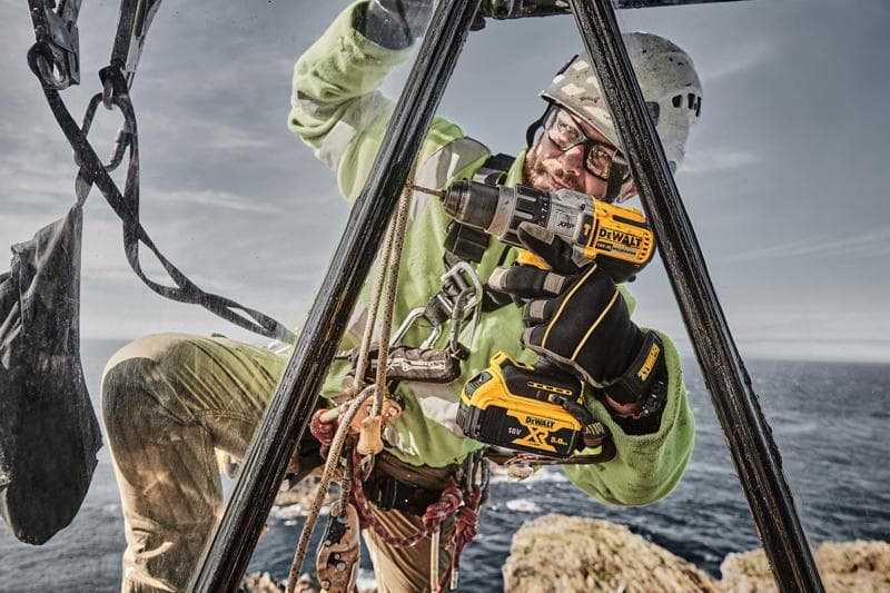 Um trabalhador equipado com equipamento de segurança utiliza uma furadeira elétrica sem fio DEWALT DCD996P2 ao ar livre, posicionado em uma estrutura metálica perto de um penhasco com o mar ao fundo.