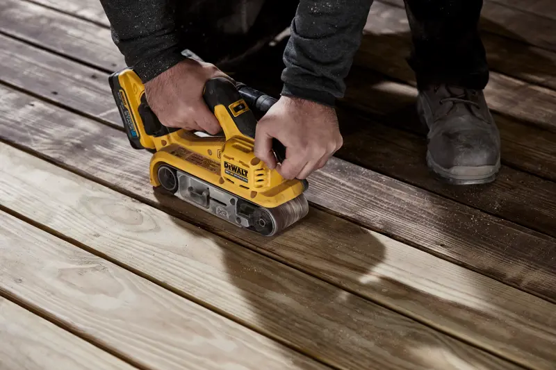 Uma pessoa utiliza uma lixadora de cinta sem fios amarela DEWALT em tábuas de madeira para alisar a superfície.
