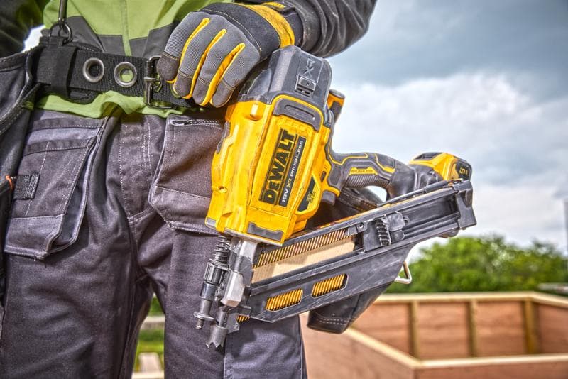Close-up de uma pessoa usando luvas de trabalho segurando uma pistola de pregos DEWALT DCN930N em um canteiro de obras, com uma estrutura de madeira ao fundo.