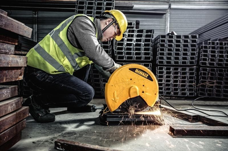 Um trabalhador usando colete de segurança e capacete utiliza uma ferramenta elétrica DEWALT D28730 para cortar metal, criando faíscas em um ambiente industrial com vigas de metal empilhadas.