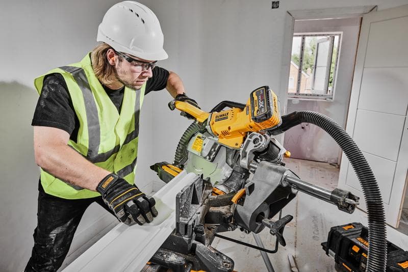 Uma pessoa usando colete de segurança, luvas e capacete está operando uma serra de esquadria sem fio DEWALT DCS781X2 para cortar um acabamento em uma sala em construção. A serra está conectada a um sistema de extração de pó, e baterias de ferramentas DEWALT estão visíveis próximas.