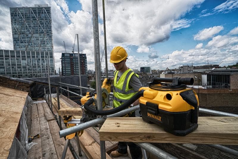 Um aspirador portátil DEWALT DCV584 em um canteiro de obras, com um trabalhador de colete refletivo e capacete operando ferramentas elétricas em um andaime, e prédios da cidade ao fundo.