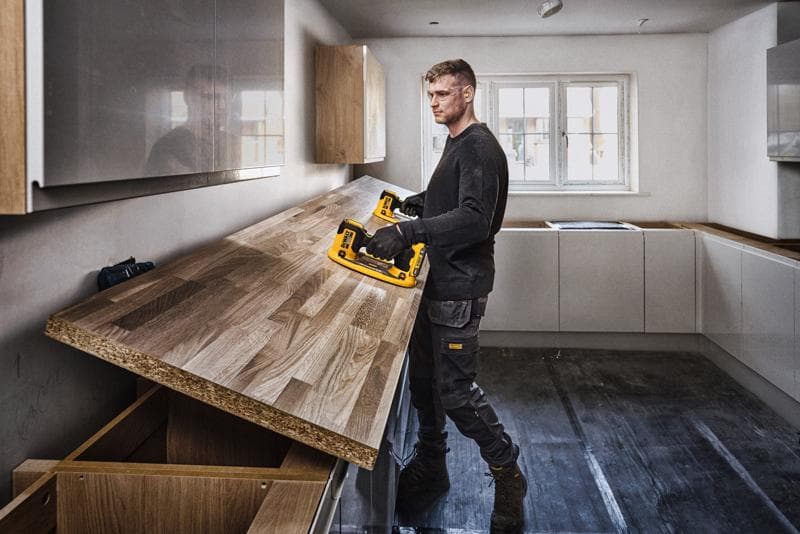 Uma pessoa com o rosto desfocado está instalando uma bancada de madeira em uma cozinha usando uma ferramenta elétrica DEWALT. A cozinha é moderna, com armários claros e uma grande janela ao fundo.