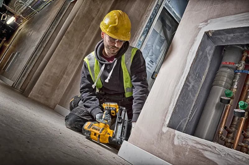 Trabalhador usa clavador DEWALT DCN660N, capacete amarelo e colete de segurança em obra.