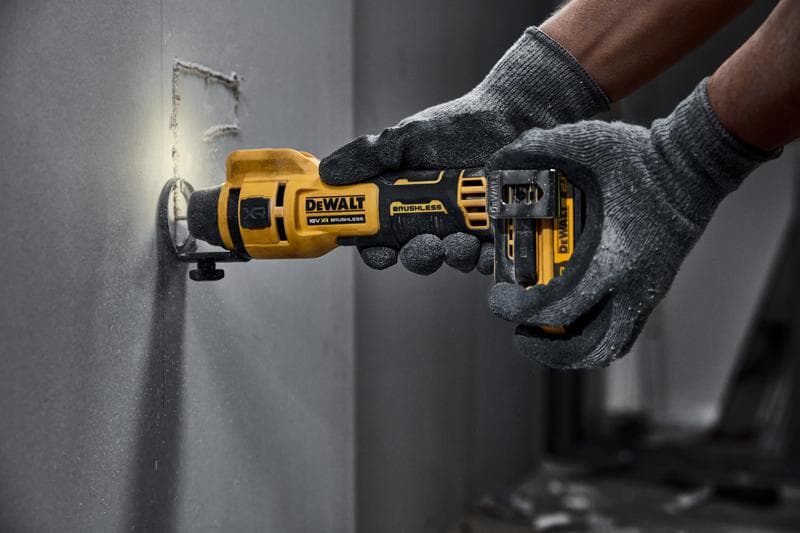 Uma pessoa usando luvas cinzas está utilizando uma ferramenta rotativa sem fio DEWALT para cortar um buraco quadrado em uma placa de drywall.