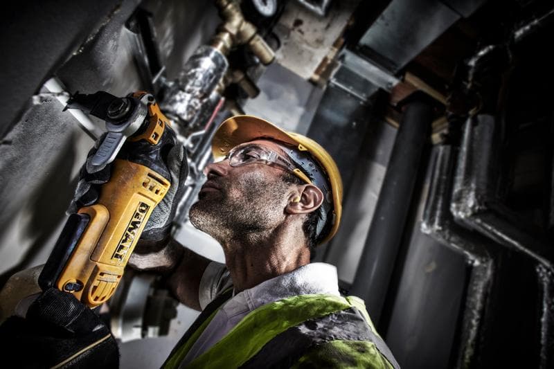 Um trabalhador com equipamento de segurança utiliza uma ferramenta elétrica DEWALT DWE315 para cortar uma parede em um ambiente industrial. O rosto do trabalhador está desfocado para proteger a privacidade.