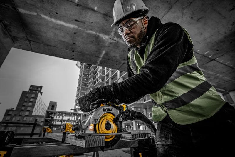 Um trabalhador da construção civil com colete de segurança e capacete opera uma serra circular DEWALT (modelo DCS565NT) para cortar uma tábua de madeira em um canteiro de obras. Ao fundo, há estruturas de concreto e prédios em construção.