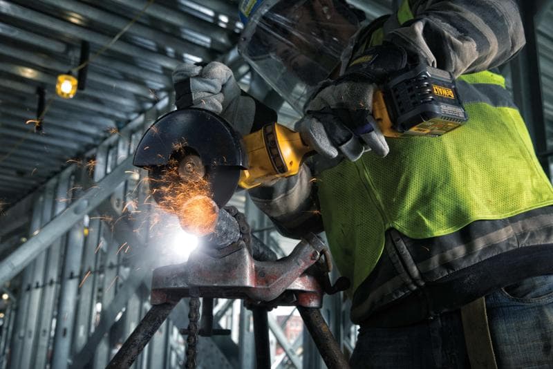 Close-up de uma pessoa usando luvas de proteção e colete de segurança operando uma esmerilhadeira angular sem fio DEWALT para cortar metal, com faíscas voando, em um local de construção industrial.