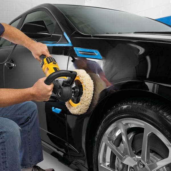 Uma pessoa utiliza uma polidora DEWALT DWP849X na parte externa preta de um carro, focando na área acima do pneu dianteiro. A polidora tem design amarelo e preto e um grande disco de polimento de lã. Fita azul está aplicada em algumas partes do carro para proteção.