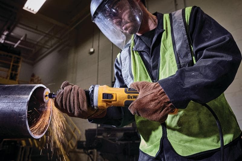 Uma pessoa usando equipamento de proteção e luvas utiliza uma esmerilhadeira angular DEWALT para cortar um tubo de metal grande, com faíscas saindo da área de corte.
