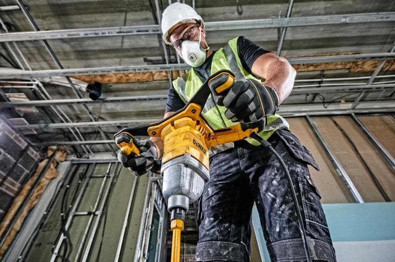 Um trabalhador da construção vestindo colete de segurança, luvas e capacete usa uma furadeira misturadora com fio amarela DEWALT (DWD241) dentro de um edifício em construção.