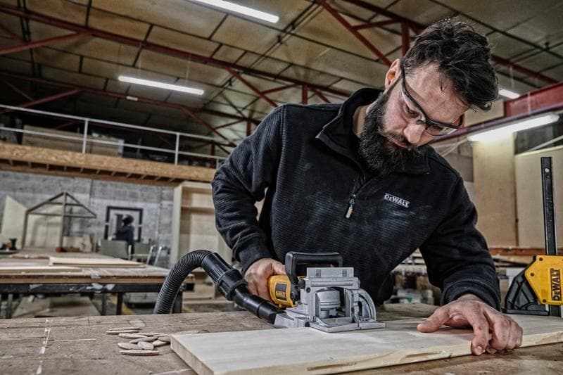 Uma pessoa usa uma ferramenta de encaixe DEWALT DW682K para trabalhar em uma peça de madeira em uma oficina. O rosto da pessoa está desfocado para preservar a privacidade. O ambiente inclui mesas, peças de madeira e equipamentos de oficina visíveis.