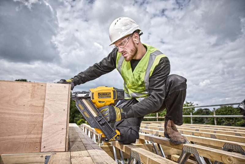 Um trabalhador da construção civil usando capacete de segurança e colete de alta visibilidade está utilizando uma pistola de pregos amarela DEWALT DCN930N para fixar tábuas de madeira em um canteiro de obras ao ar livre. O rosto do trabalhador está desfocado para proteger a privacidade.