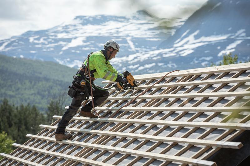 Um trabalhador da construção civil usando equipamento de segurança utiliza uma pregadora sem fio DEWALT para fixar ripas de madeira em um telhado inclinado, com uma paisagem de montanha ao fundo.