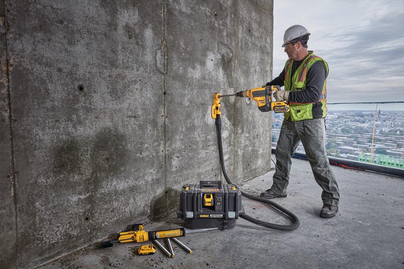 Um trabalhador da construção civil usando colete de segurança e capacete está utilizando uma furadeira sem fio DEWALT com acessório de extração de poeira para perfurar uma parede de concreto. O trabalhador está em um grande edifício com vista para a cidade ao fundo. Várias ferramentas DEWALT e uma caixa de ferramentas estão no chão próximas.