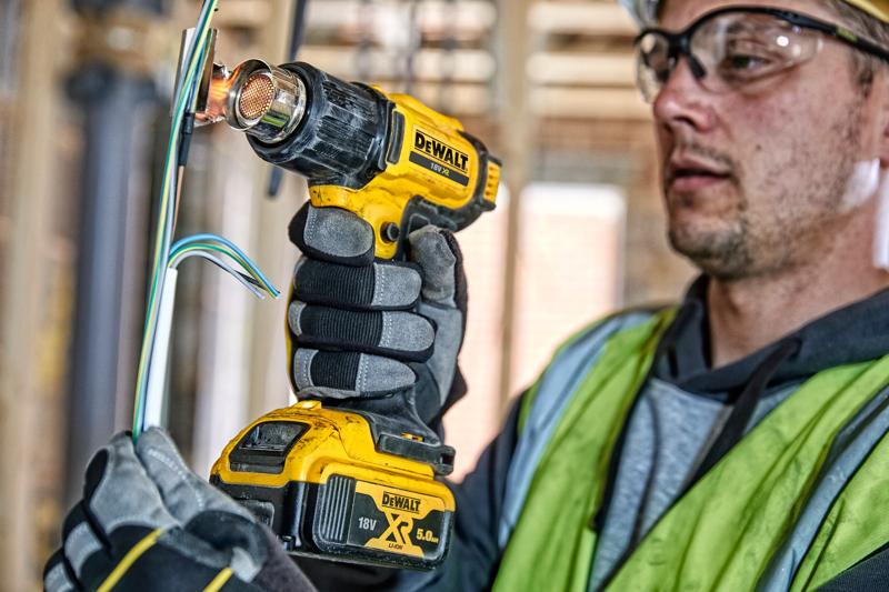 Uma pessoa usando luvas e colete de segurança está utilizando uma pistola de calor sem fio DEWALT 18V XR para trabalhar em fios elétricos em um canteiro de obras. O rosto da pessoa está desfocado.