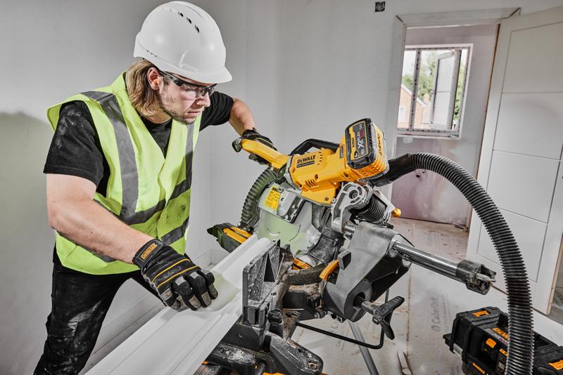 Uma pessoa usando colete de segurança, luvas e capacete está operando uma serra de esquadria sem fio DEWALT DCS781X2 para cortar um acabamento em uma sala em construção. A serra está conectada a um sistema de extração de pó, e baterias de ferramentas DEWALT estão visíveis próximas.