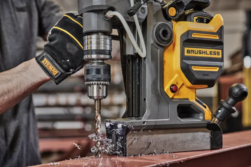 Pessoa fura metal com broca magnética DEWALT brushless, gerando cavacos de metal na mesa de oficina.