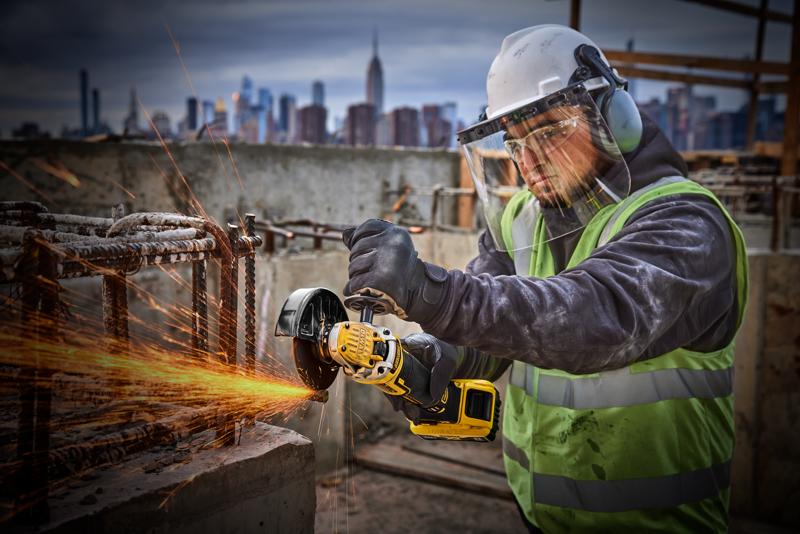 Uma pessoa usando equipamentos de proteção, incluindo capacete, viseira, luvas e colete refletivo, está usando uma esmerilhadeira angular sem fio DEWALT amarela (SKU: DCG405P2) para cortar vergalhões de metal em um canteiro de obras. Faíscas saem da área de corte. O horizonte da cidade está visível ao fundo.