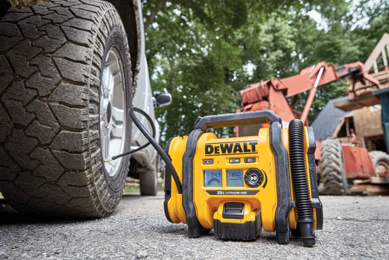 Um inflador de pneus portátil DEWALT está sendo usado, conectado ao pneu de um veículo, em um local de trabalho ao ar livre com equipamentos de construção ao fundo.