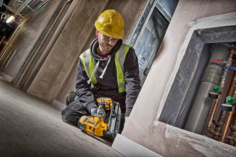 Trabalhador usa clavador DEWALT DCN660N, capacete amarelo e colete de segurança em obra.