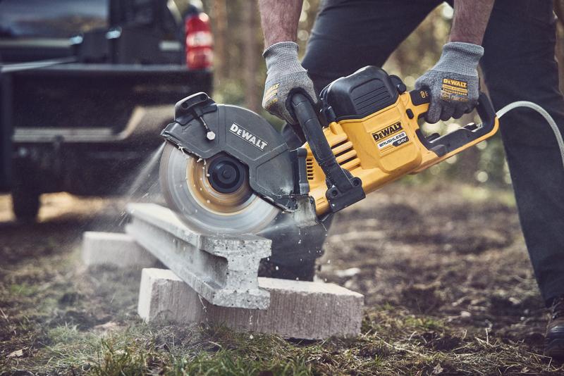Uma pessoa usando luvas está utilizando uma serra elétrica sem fio DEWALT para cortar uma laje de concreto ao ar livre. A ferramenta é amarela e preta com a marca visível, e poeira de concreto é vista enquanto a serra está em ação.