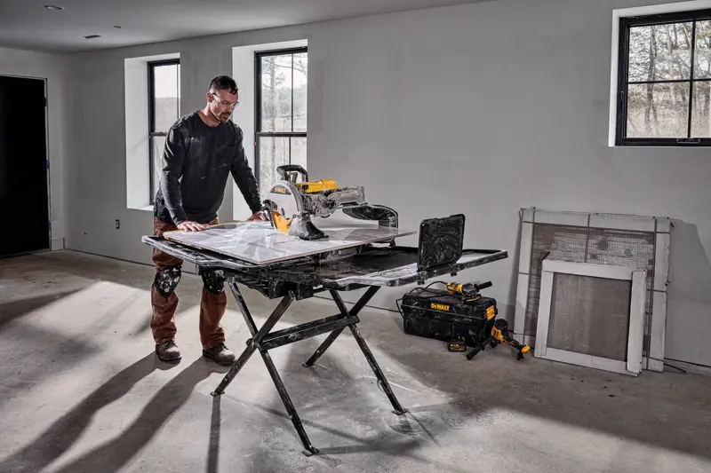 Uma pessoa está ao lado de uma serra para azulejos DEWALT D36000S montada em seu suporte em uma sala em construção. A serra está em um suporte preto dobrável, e há ferramentas e uma bolsa de ferramentas DEWALT no chão próximo. A sala possui paredes inacabadas, janelas e materiais de construção encostados na parede.