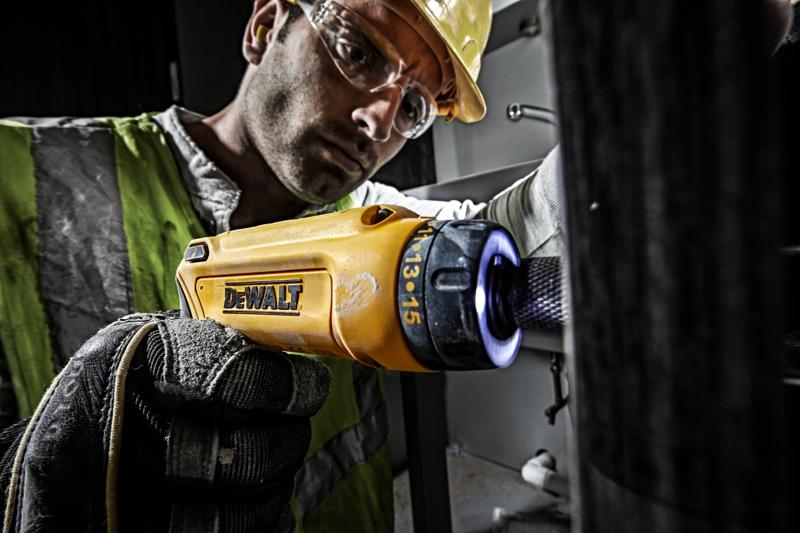 Uma pessoa usando colete de segurança refletivo e luvas utiliza uma chave de fenda sem fio amarela DEWALT (modelo DCF680G2) para trabalhos de manutenção internos. O foco está na ferramenta e na mão enluvada do usuário. O rosto da pessoa está borrado por motivos de privacidade.