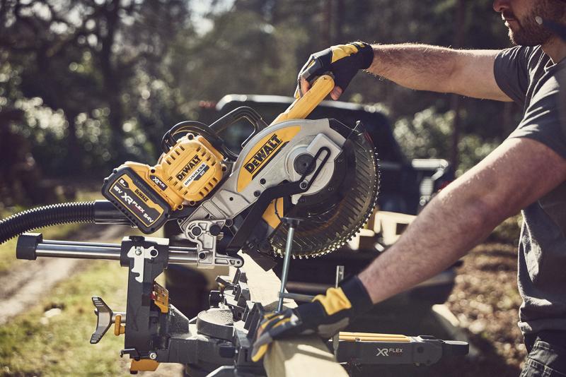 Uma pessoa usando luvas opera uma serra de esquadria deslizante sem fio DEWALT ao ar livre para cortar um pedaço de madeira. A serra tem design amarelo e preto com a marca DEWALT XR FLEXVOLT.