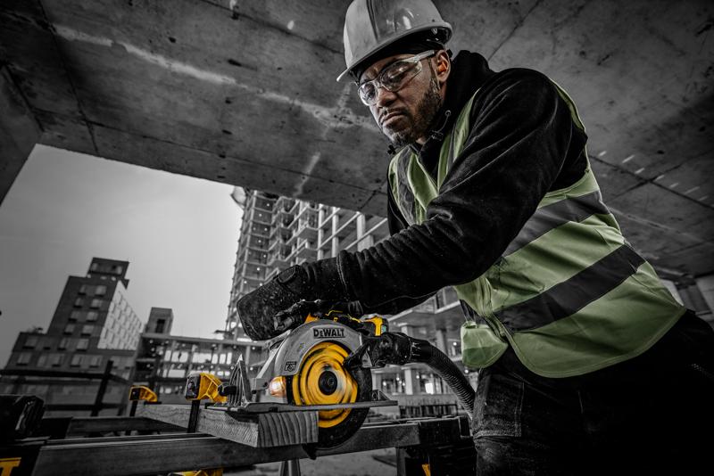Um trabalhador da construção civil com colete de segurança e capacete opera uma serra circular DEWALT (modelo DCS565NT) para cortar uma tábua de madeira em um canteiro de obras. Ao fundo, há estruturas de concreto e prédios em construção.