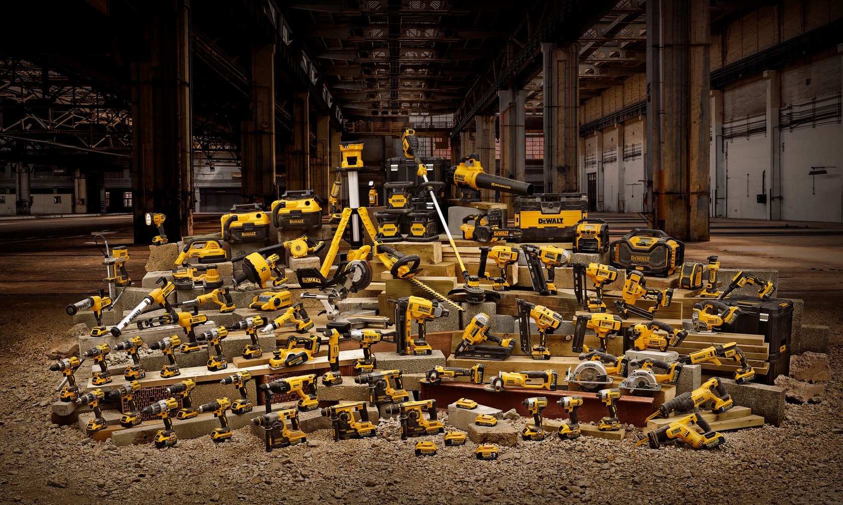 Uma grande coleção de ferramentas elétricas, acessórios e caixas de ferramentas DEWALT organizadas sobre blocos de concreto em um ambiente de armazém industrial. As ferramentas incluem furadeiras, serras, esmerilhadeiras e outros equipamentos de construção, todos nas cores amarelo e preto distintivas da DEWALT.